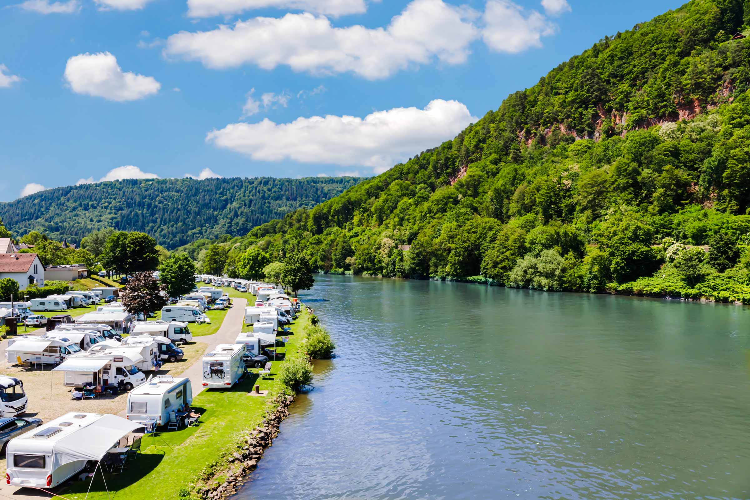 Ein Fluss mit grünen Ufern, Campingfahrzeugen und bewaldeten Bergen unter blauem Himmel mit Wolken.